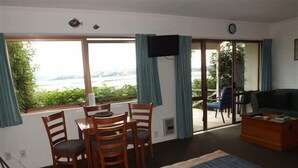 Una habitación con cama, televisor, mesa con sillas y vistas a zonas verdes y un cuerpo de agua a través de amplias ventanas.