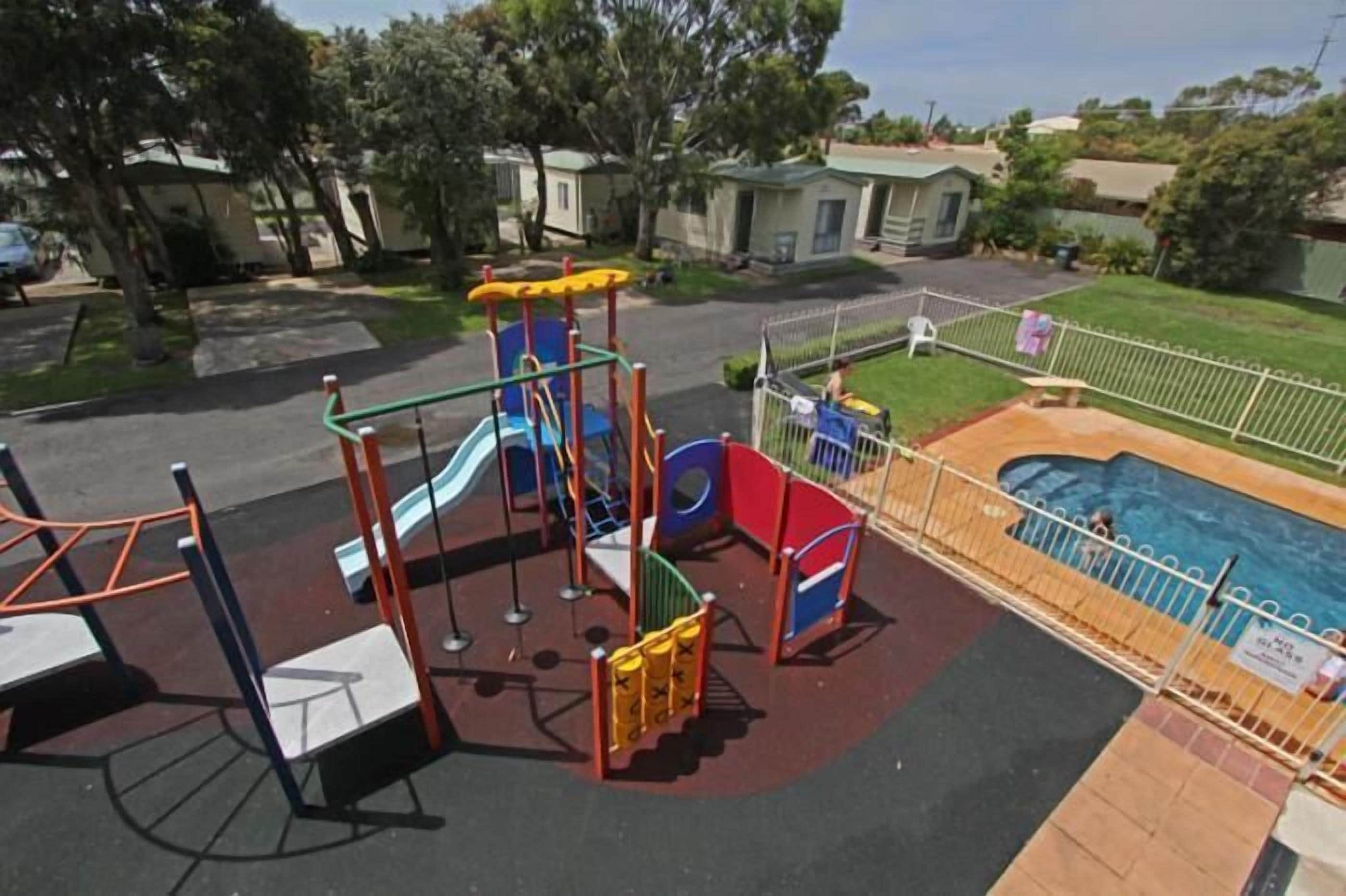 Un parque infantil con toboganes y estructuras para escalar, una piscina vallada y una zona ajardinada con árboles.