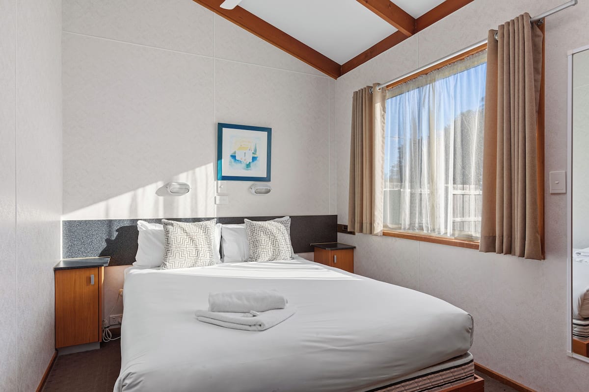 A hotel room with a large bed, a nightstand, a wall-mounted lamp, a window with curtains, and a framed picture on the wall.