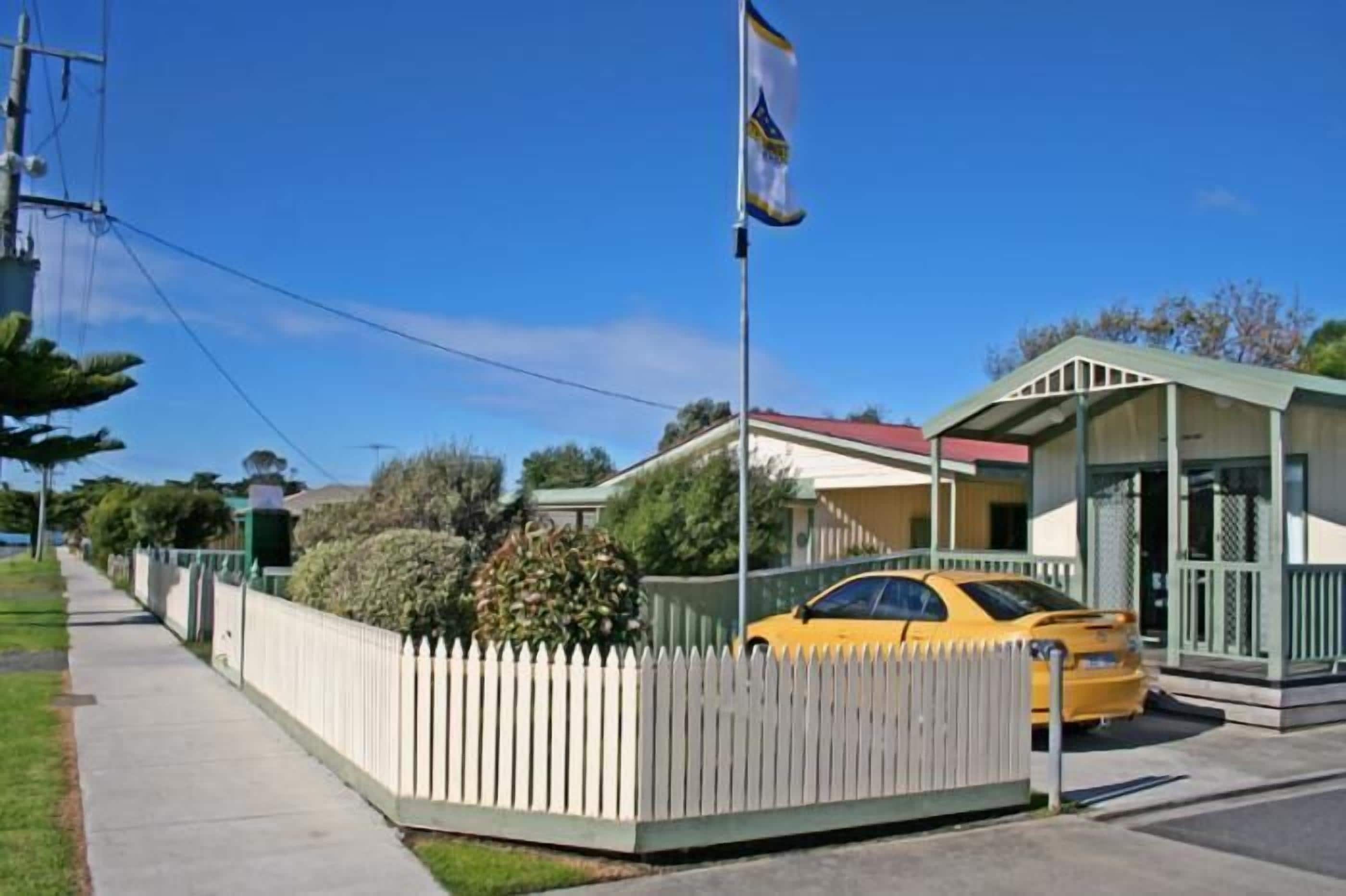 A single-story building with a red roof, a white picket fence, and a yellow car parked in front.