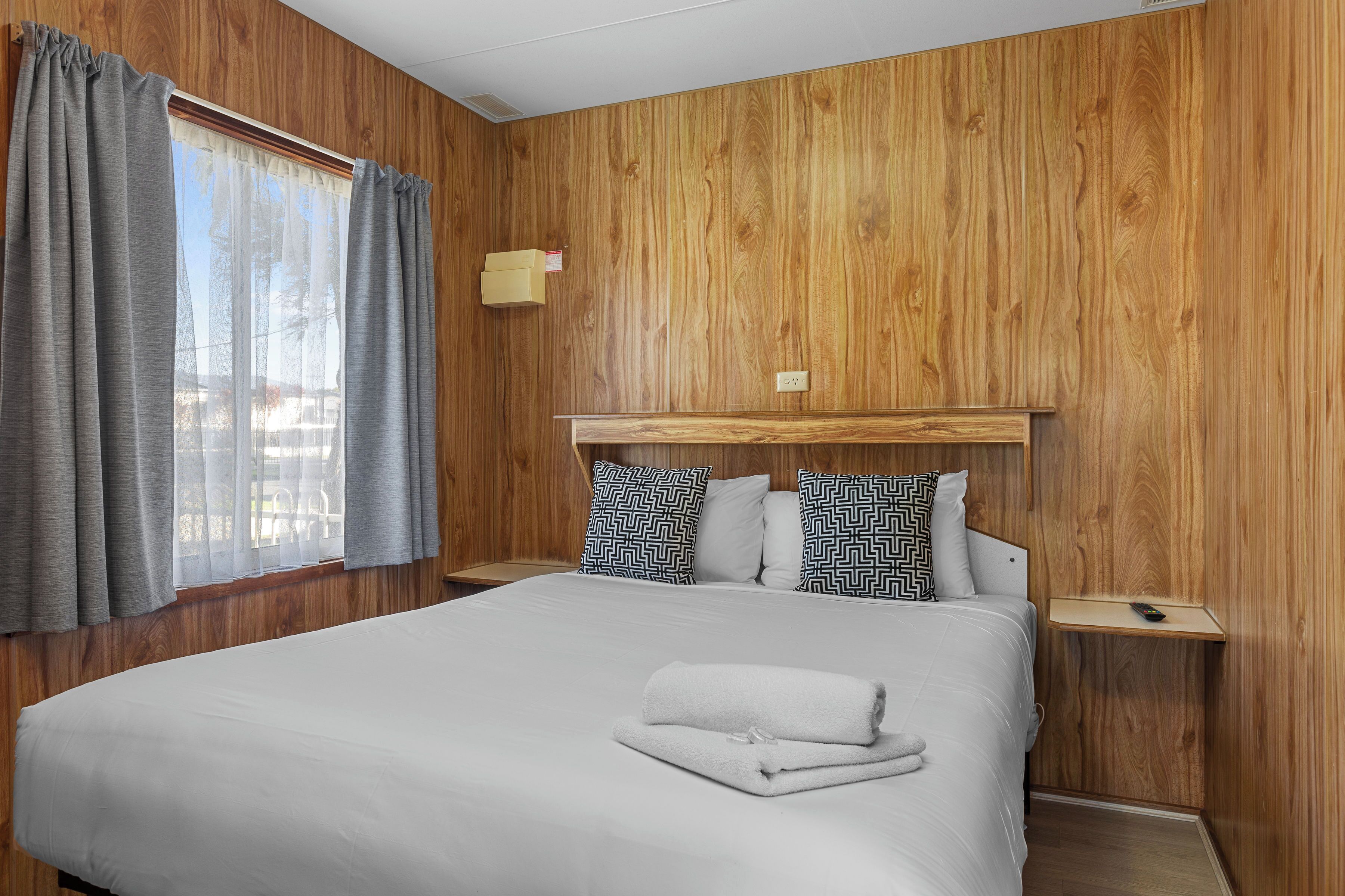 Ein Hotelzimmer mit einer holzverkleideten Wand, einem Bett mit weißer Bettwäsche, grauen Vorhängen und einem Nachttisch mit einer Lampe.