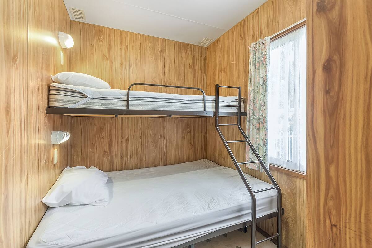 A bunk bed room with wooden walls, a window with floral curtains, and a wall-mounted light fixture.