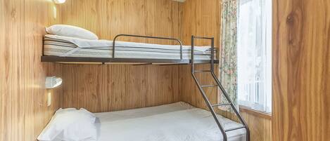 A bunk bed room with wooden walls, a window with floral curtains, and a wall-mounted light fixture.
