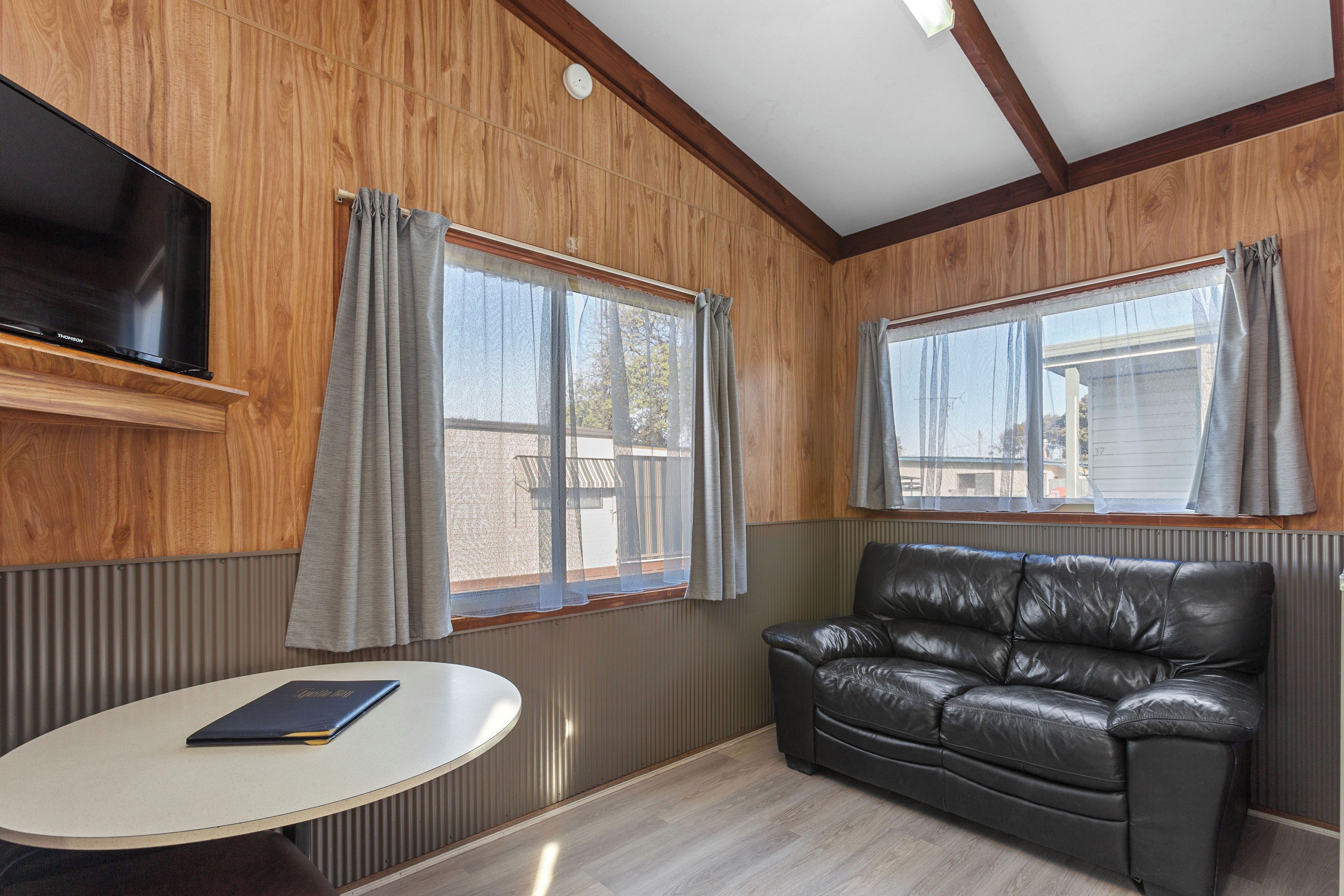 A room with a TV mounted on the wall, a leather sofa, a round table, and two windows with curtains.