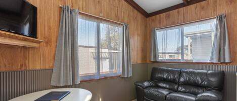 A room with a TV mounted on the wall, a leather sofa, a round table, and two windows with curtains.