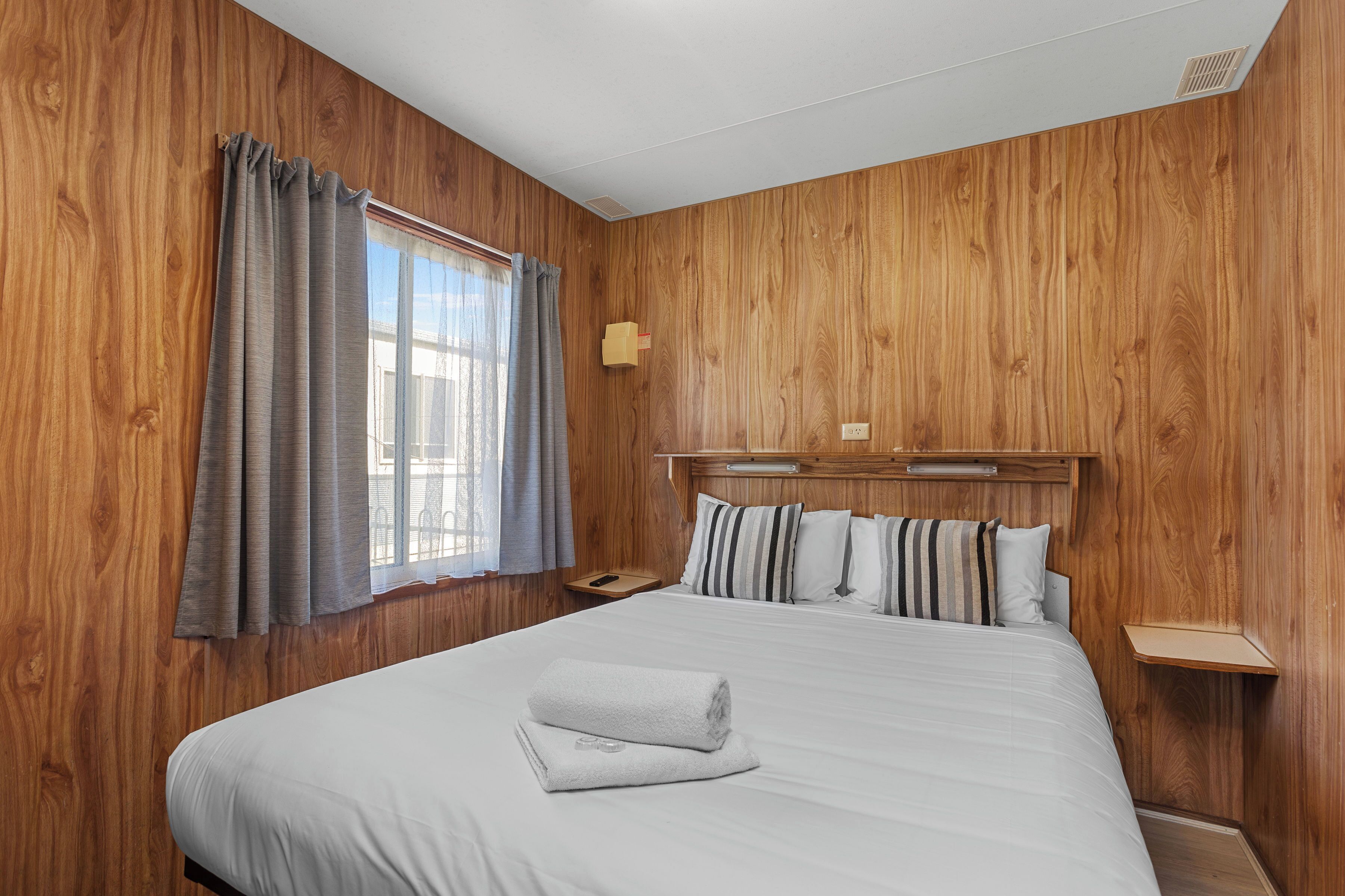 A hotel room with wooden panel walls, a large bed with white bedding, striped pillows, a bedside table, and a window with curtains.