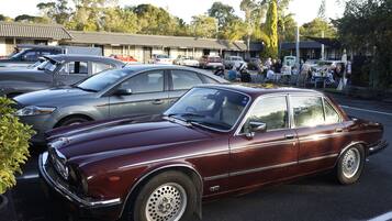 A vintage car parked in a lot with other vehicles, surrounded by trees and buildings in the background.