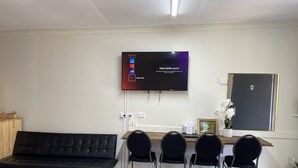 Una habitación moderna con una televisión de pantalla plana en la pared, un sofá negro, un escritorio de madera con un cuadro enmarcado y cuatro sillas negras.
