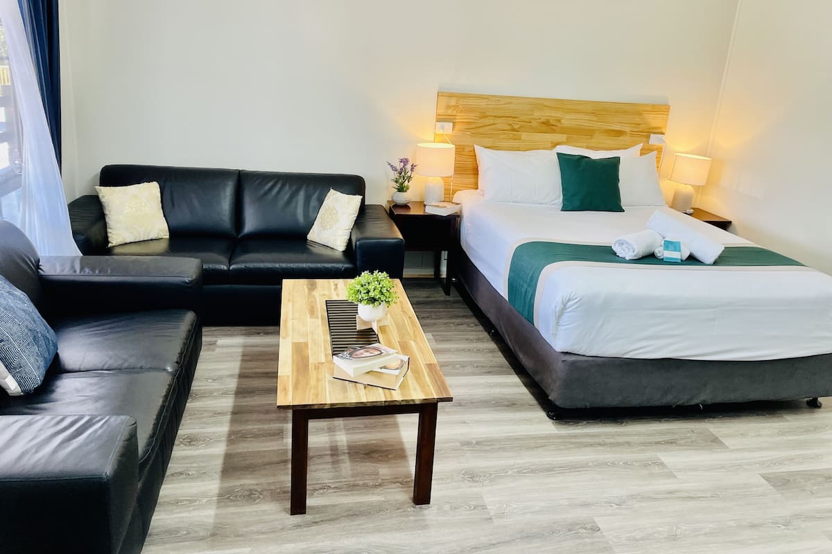A hotel room with a black leather sofa, a wooden coffee table with a plant, a bed with white bedding and green pillows, and two bedside lamps.