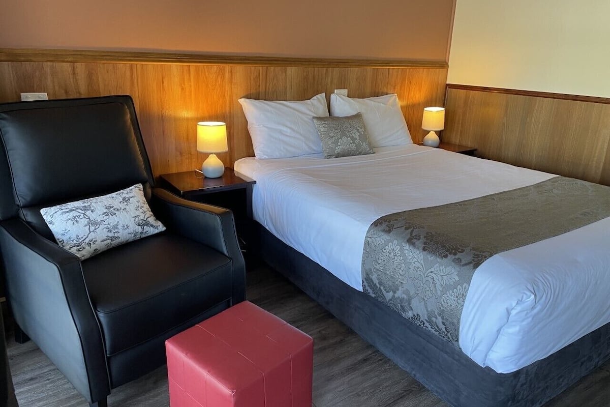 A hotel room with a bed, a chair, a nightstand, and a red ottoman.