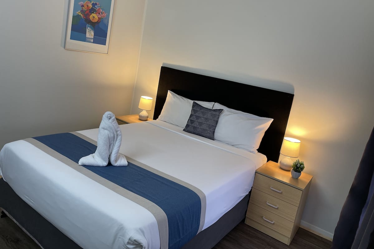 A neatly made bed with a blue and white bedspread, two pillows, a white towel, and a bedside lamp on a wooden nightstand.