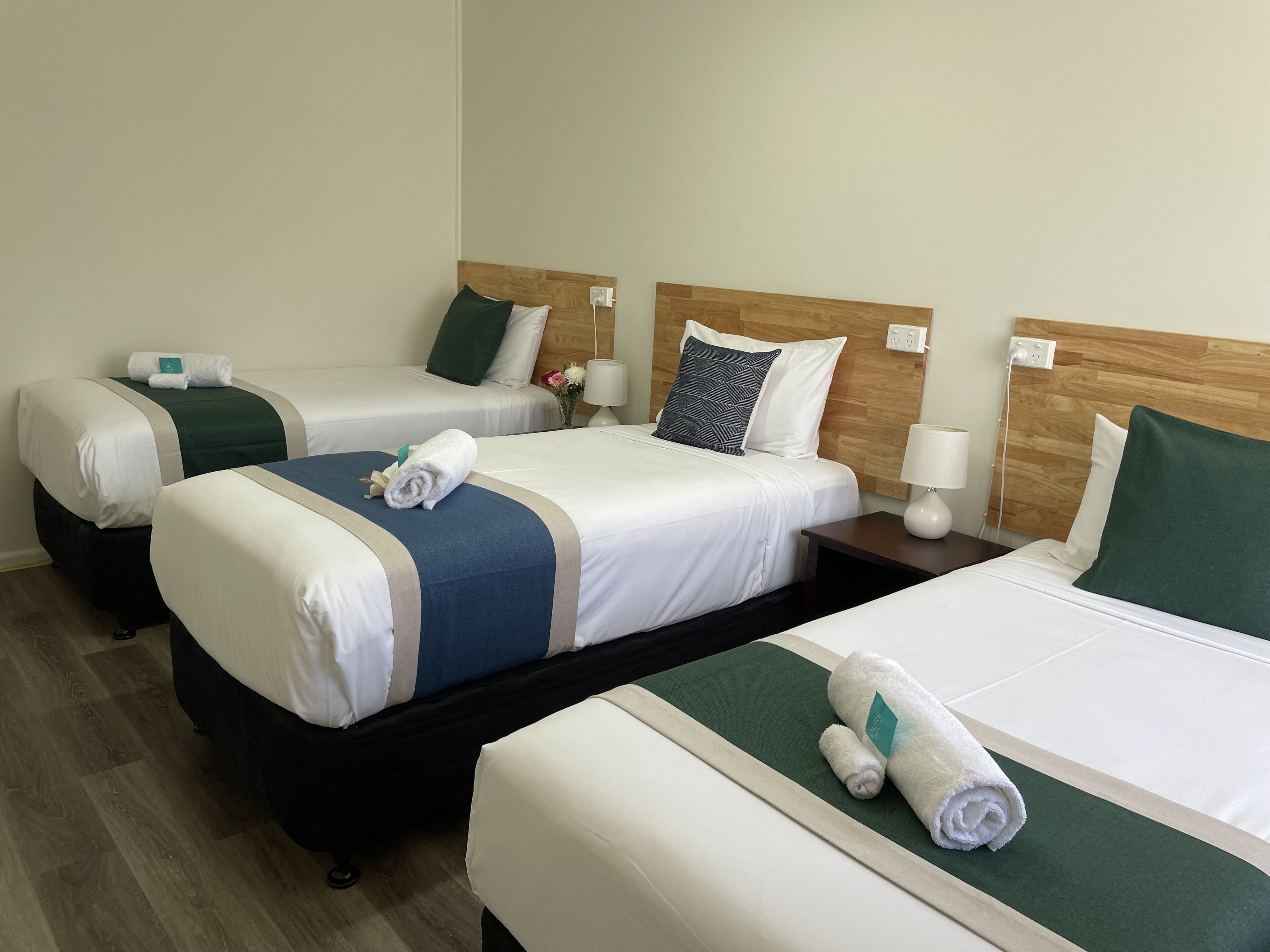 Habitación de hotel con tres camas, cada una con cabecero de madera, ropa de cama blanca y detalles en verde. Se observan mesitas de noche con lámparas y un pequeño jarrón con flores sobre una de las camas.