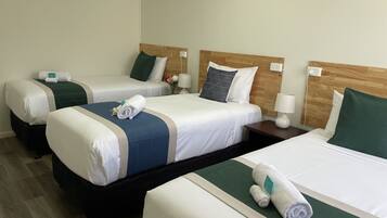 Habitación de hotel con tres camas, cada una con cabecero de madera, ropa de cama blanca y detalles en verde. Se observan mesitas de noche con lámparas y un pequeño jarrón con flores sobre una de las camas.