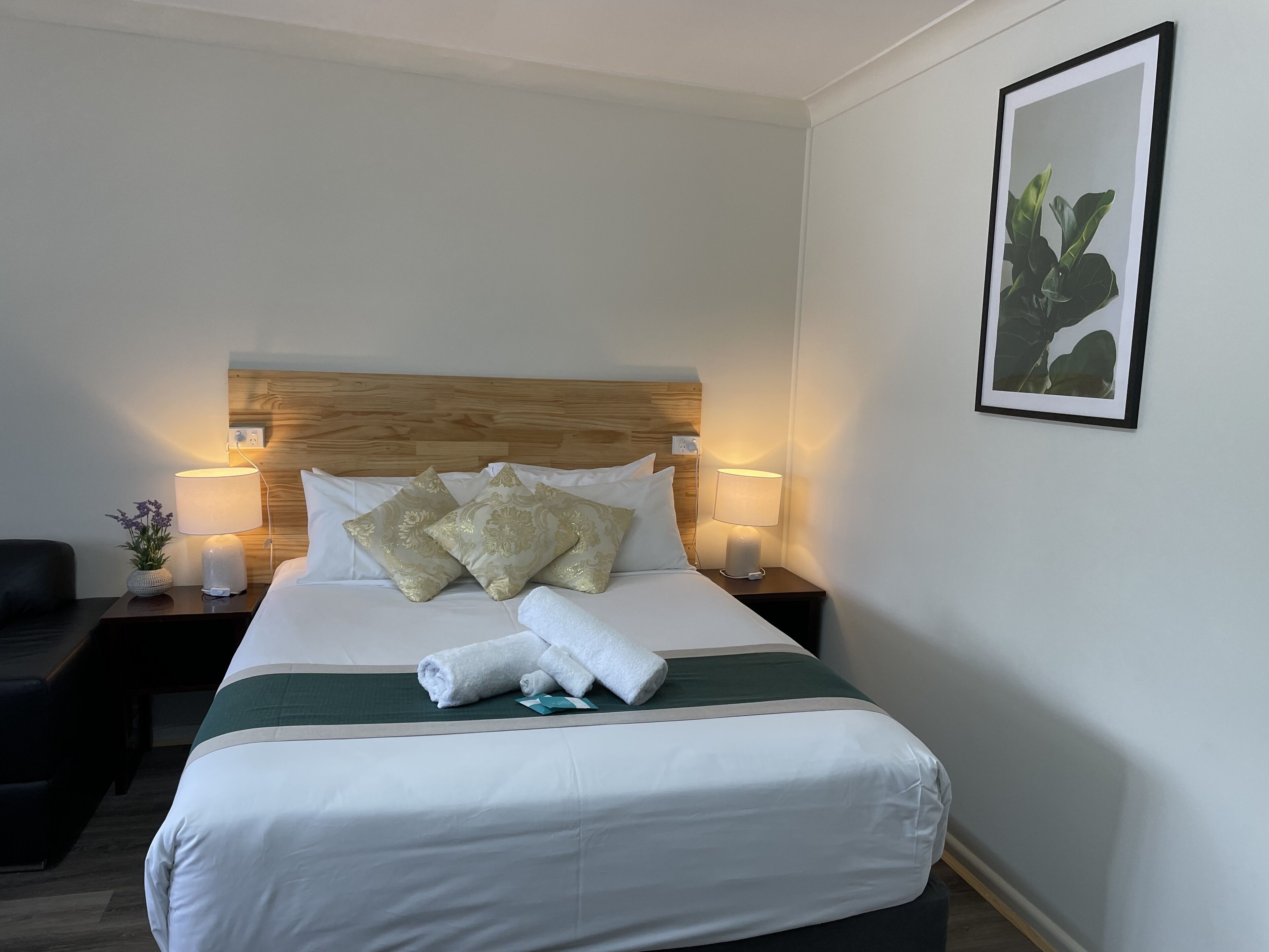 A neatly made bed with white and green bedding, two decorative pillows, a headboard, bedside lamps, a small table with a vase, and a framed picture on the wall.