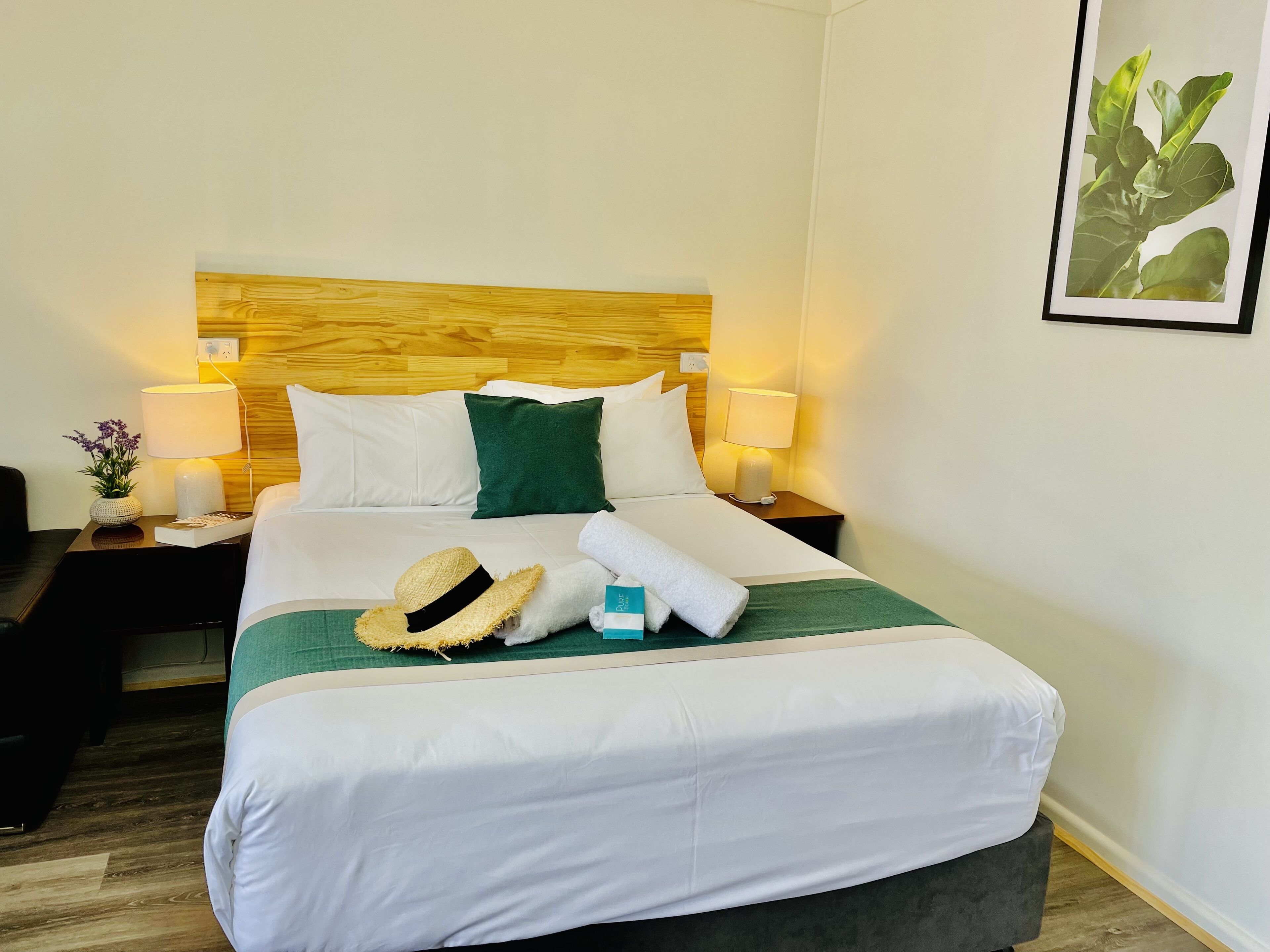 A neatly made bed with white linens, a green pillow, and a straw hat on the bed. There are two bedside lamps on wooden nightstands, one with a vase of flowers. A framed picture hangs on the wall to the right.