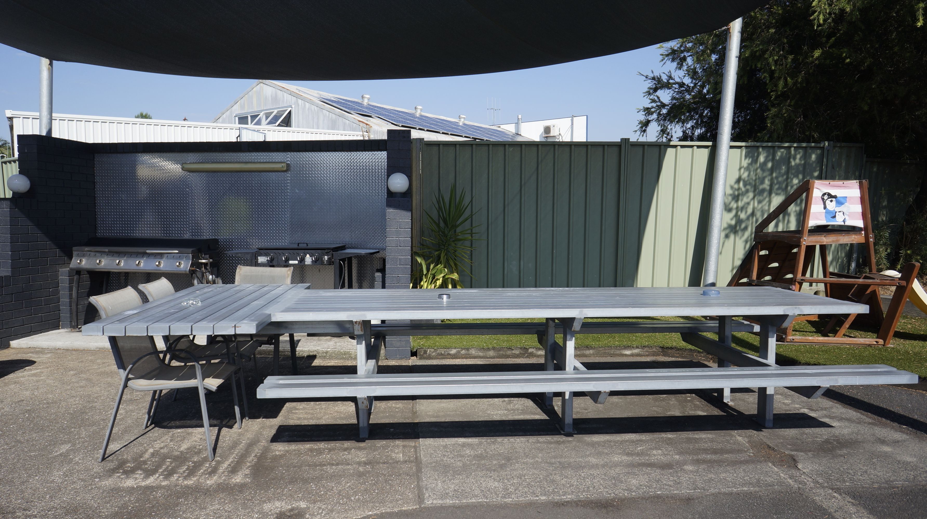 Un patio al aire libre con mesa y sillas de metal, una parrilla y una zona con sombra.