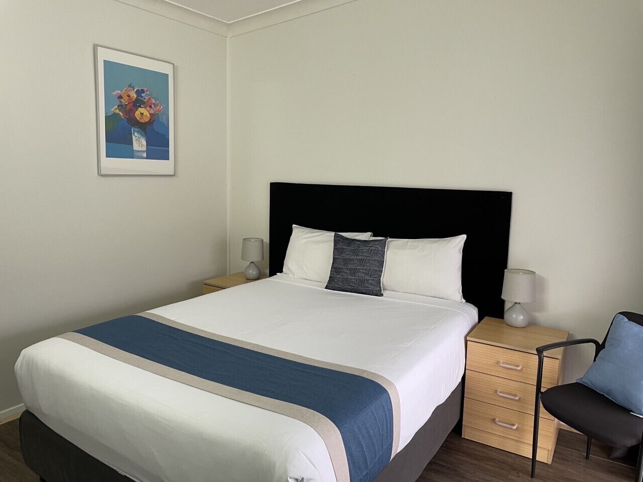 Una cama bien hecha con un detalle azul, un cabecero negro y dos mesitas de noche con lámparas.