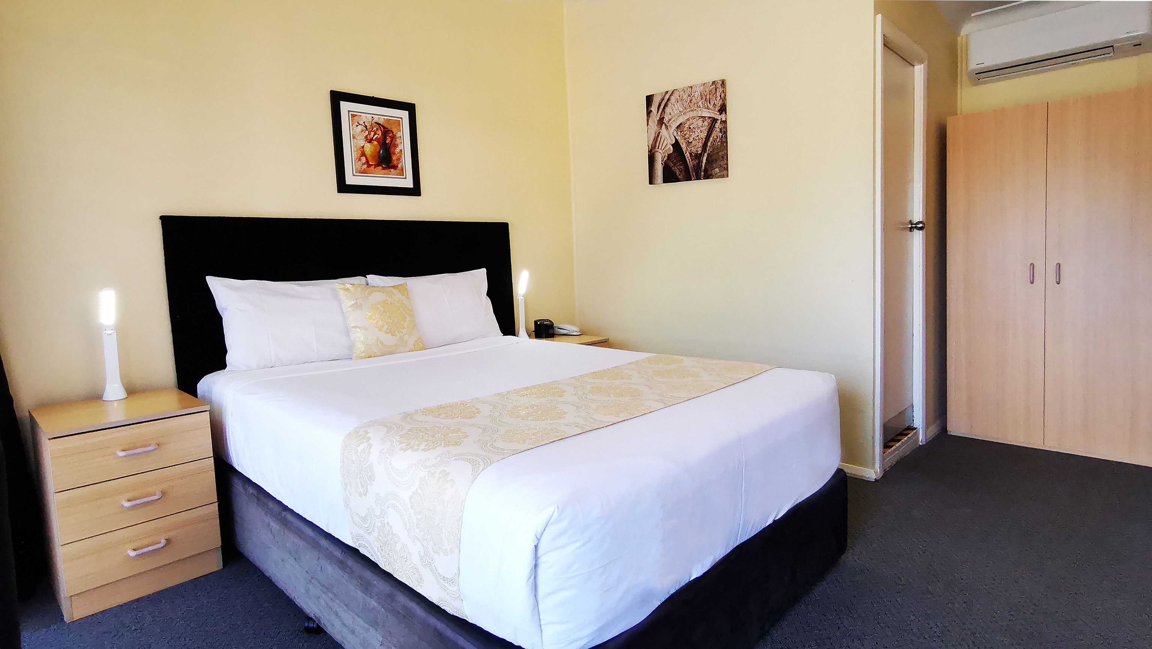 A neatly made bed with white bedding and a decorative runner, a bedside lamp, a nightstand with drawers, and two framed pictures on the wall.
