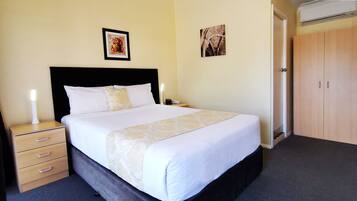A neatly made bed with white bedding and a decorative runner, a bedside lamp, a nightstand with drawers, and two framed pictures on the wall.