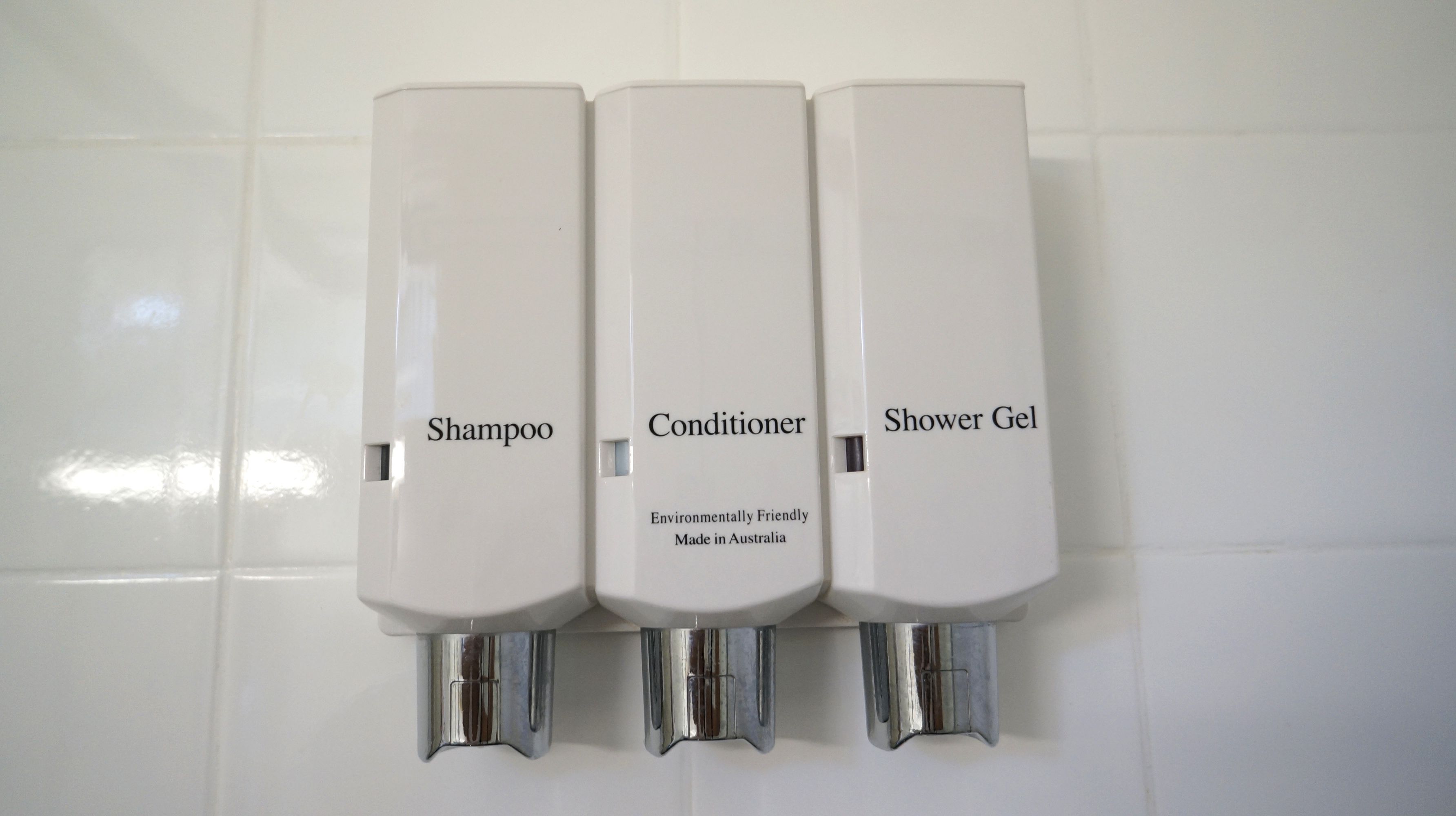 Three white dispensers labeled 'Shampoo,' 'Conditioner,' and 'Shower Gel' mounted on a tiled wall.