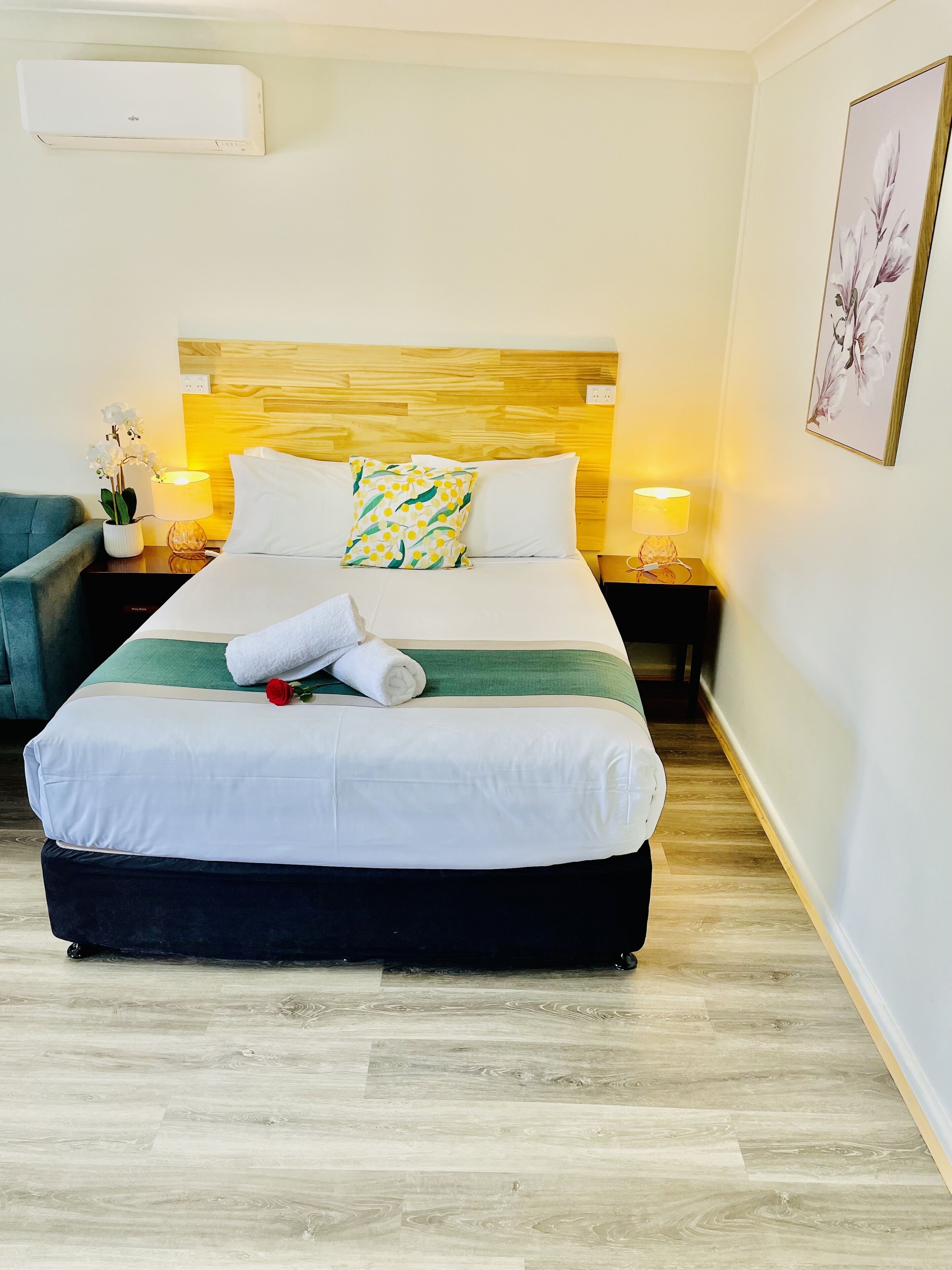 Una cama bien hecha con una manta verde y blanca a rayas, una almohada blanca y una toalla blanca enrollada. Hay una rosa roja sobre la cama. La habitación tiene un cabecero de madera, una mesita de noche con una lámpara y un cuadro en la pared.