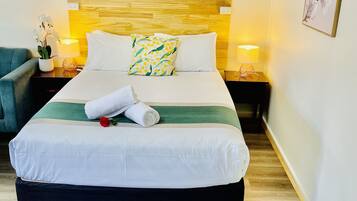 A neatly made bed with a green and white striped blanket, a white pillow, and a rolled white towel. There is a red rose on the bed. The room has a wooden headboard, a nightstand with a lamp, and a framed picture on the wall.