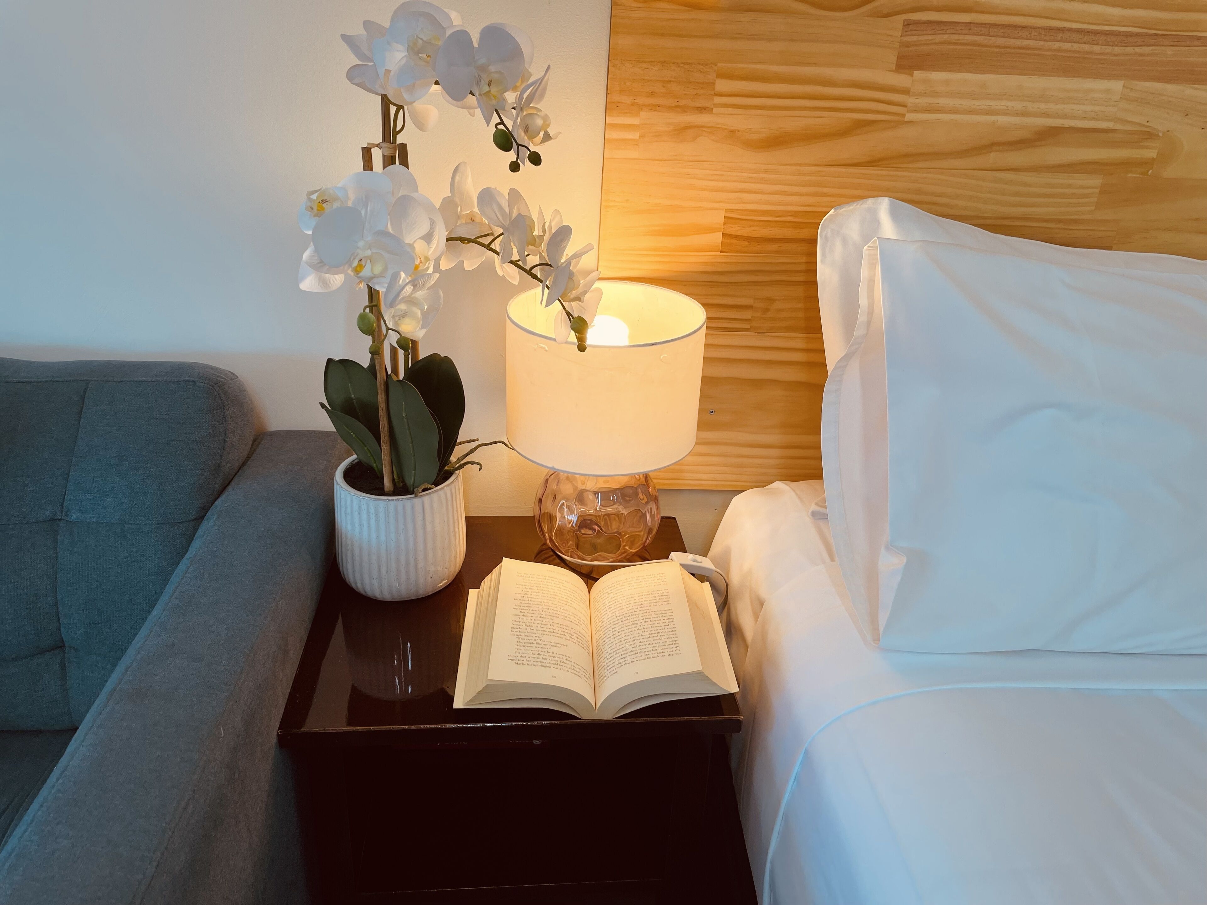 A neatly made bed with white linens, a bedside table with a lamp, a book, and a potted orchid plant.