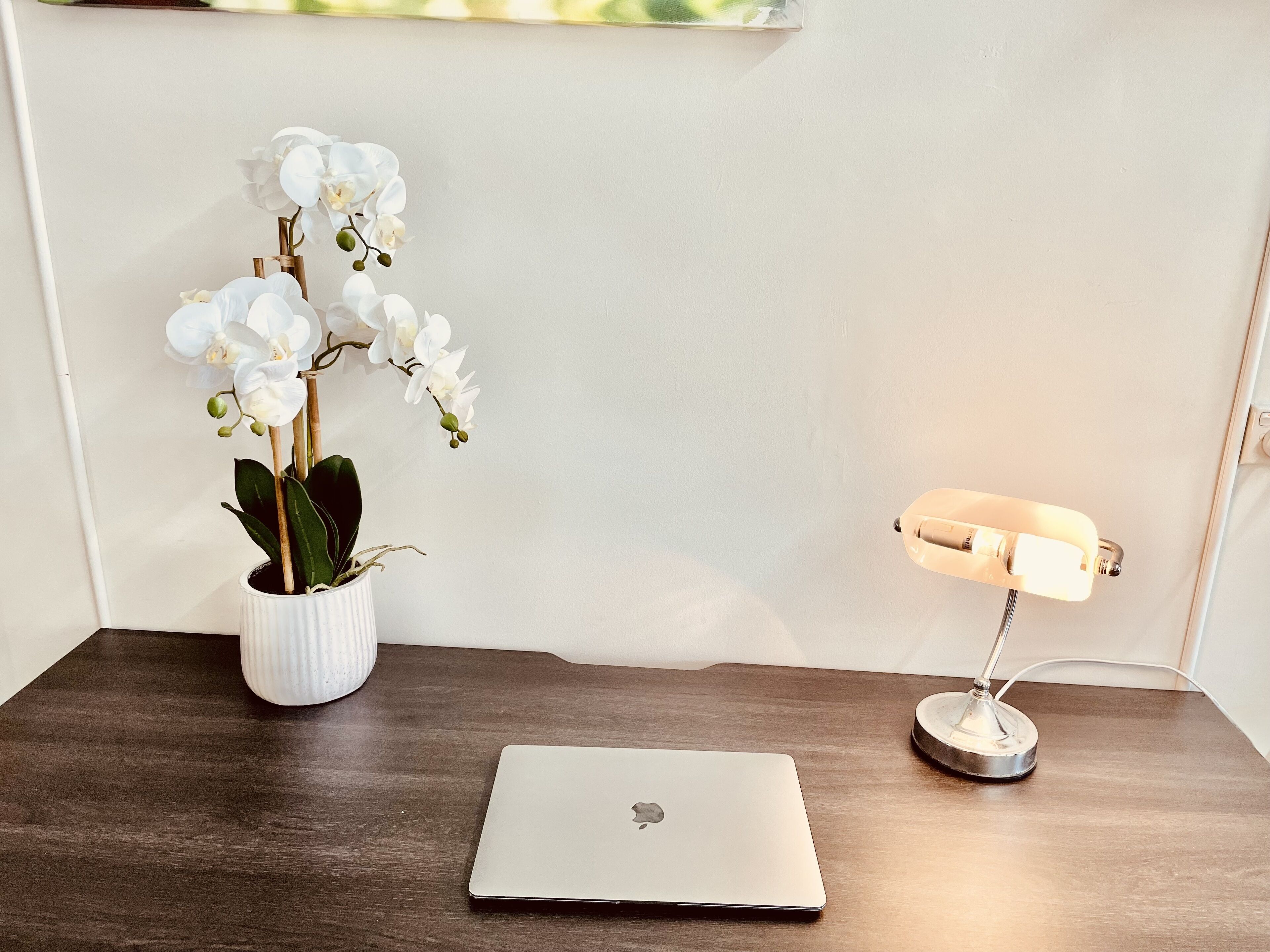 A laptop on a wooden desk, a white vase with white orchids, and a desk lamp.