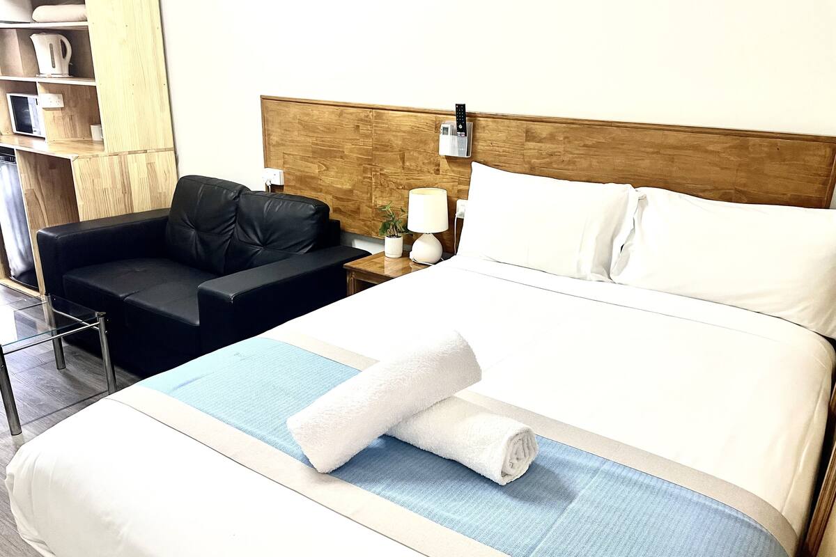 A neatly made bed with white linens and a rolled towel, a black leather sofa, a bedside table with a lamp, and a wooden headboard.
