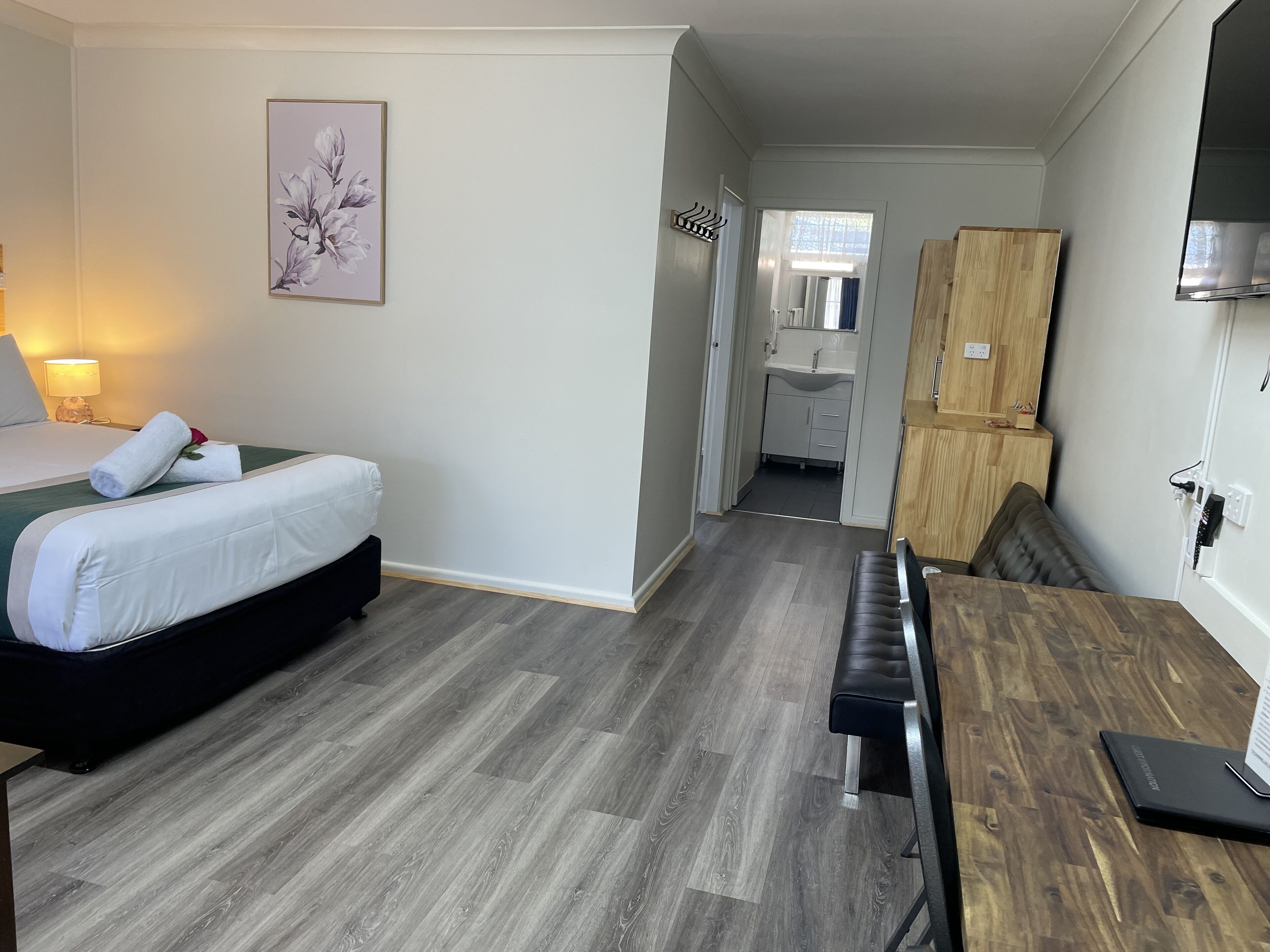 Ein Hotelzimmer mit einem Bett, einer Holzbank, einem Schreibtisch, einem Spiegel, einem Bild und Blick ins Bad.