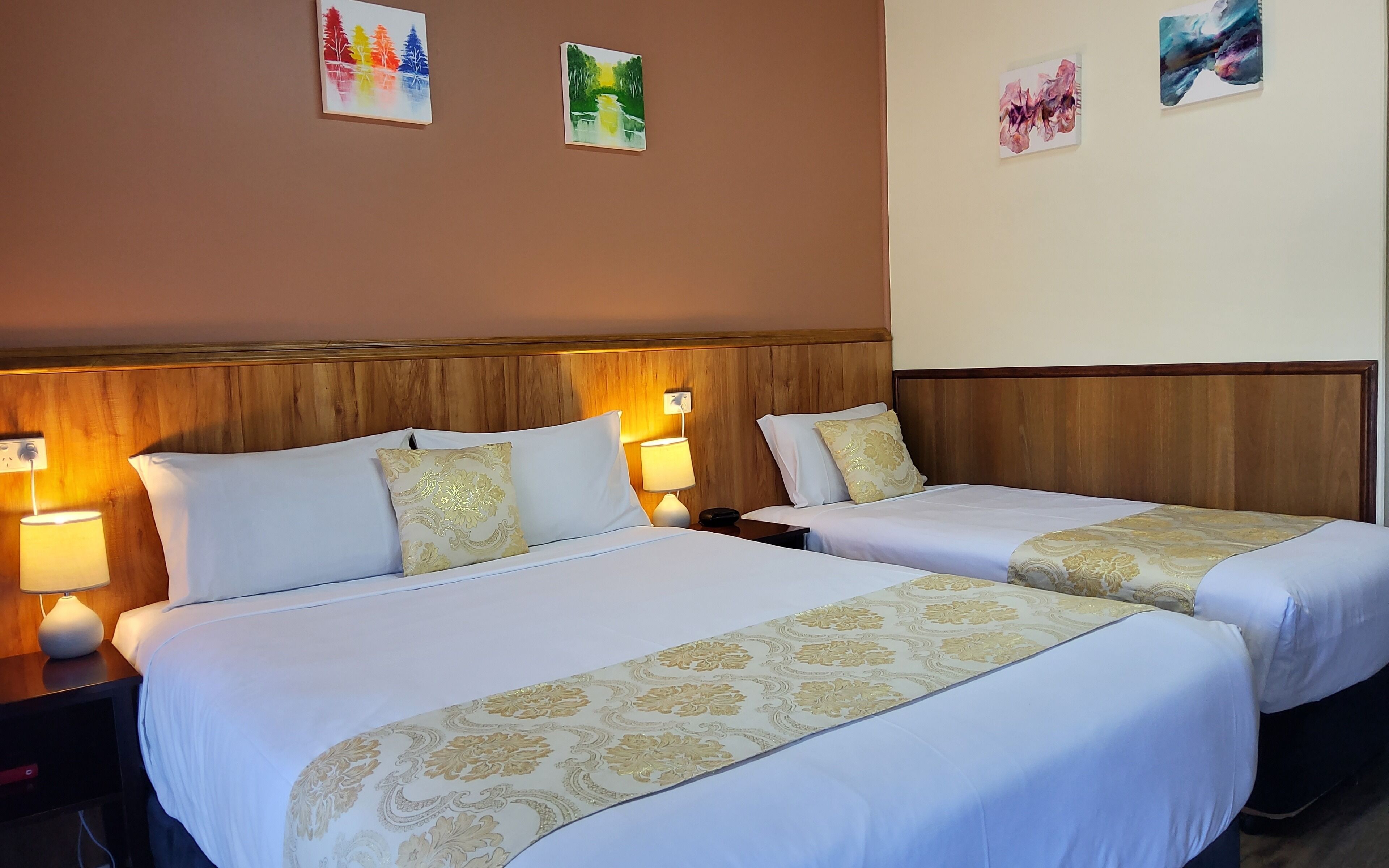 A hotel room with two beds, each with a white bedspread and gold trim, flanked by nightstands with lamps.