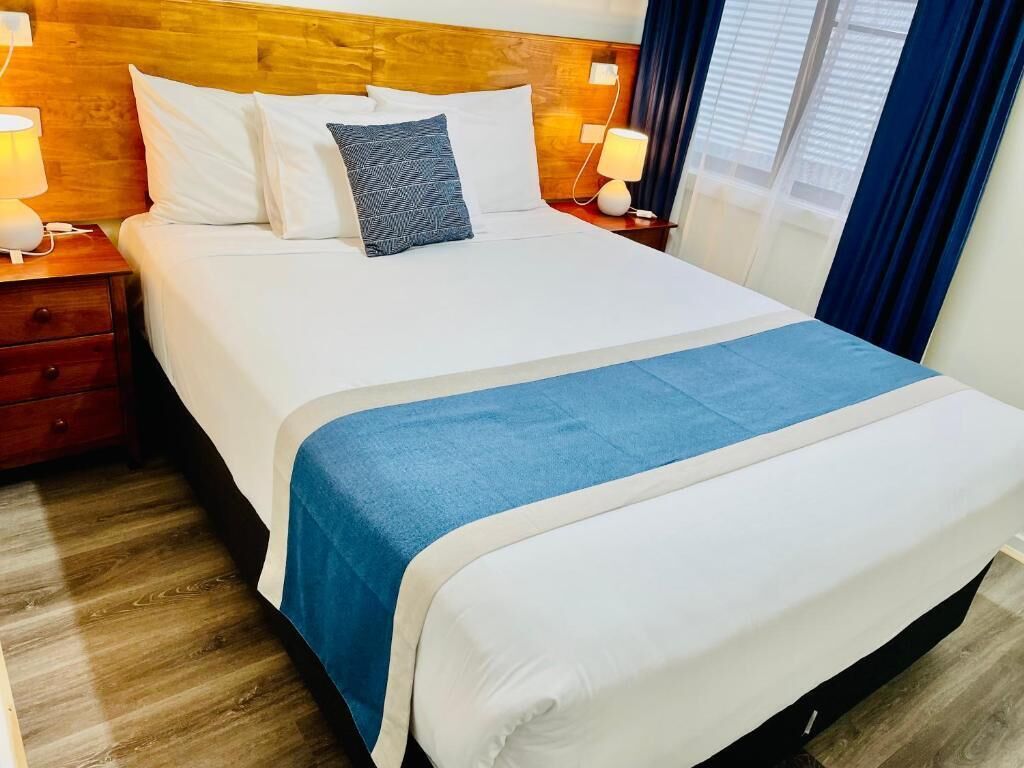 Un lit bien fait, avec du linge de lit blanc et un plaid bleu, une tête de lit en bois, des lampes de chevet et une table de chevet avec un tiroir.