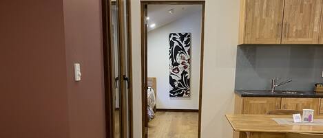 A hotel room with a bed, a wooden desk, a kitchenette, and a door leading to another room.
