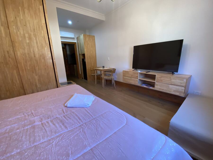 A bedroom with a large bed, a TV on a wooden stand, a small table with chairs, and a wardrobe.