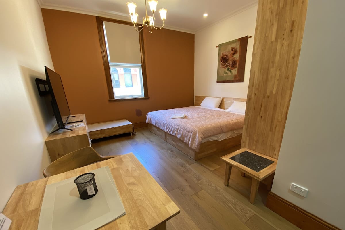 A bedroom with a bed, a desk, a chair, a small table, a TV, and a window.