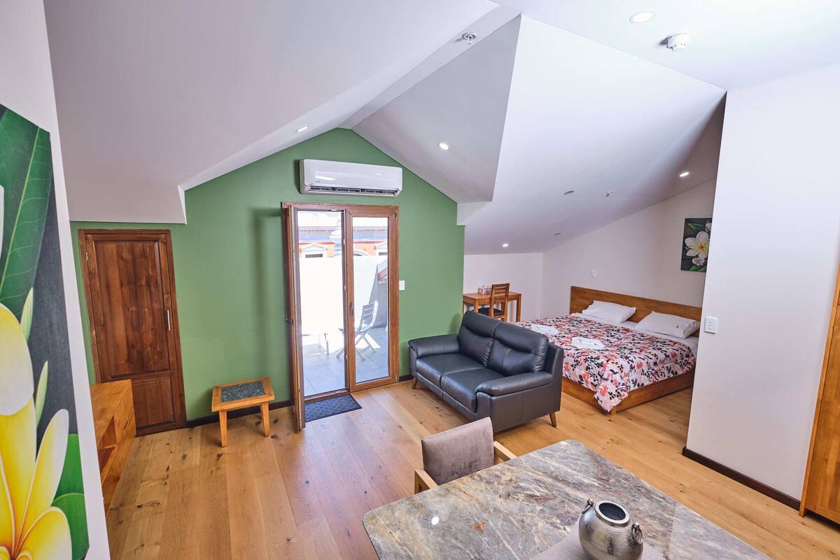 A room with a bed, a sofa, a dining table, and a small stool. There is a wooden door and a window with a view outside.
