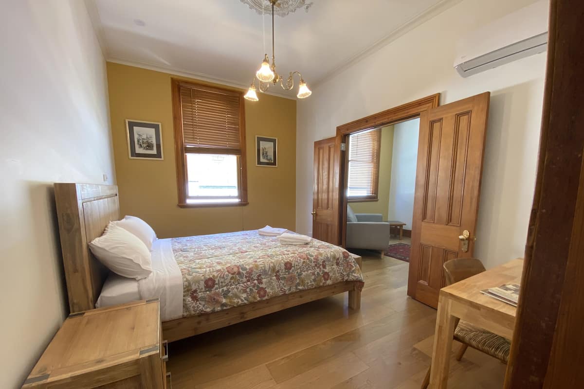 A bedroom with a wooden bed, a desk, and a chair. There are two windows with blinds, a ceiling light, and a door leading to another room.