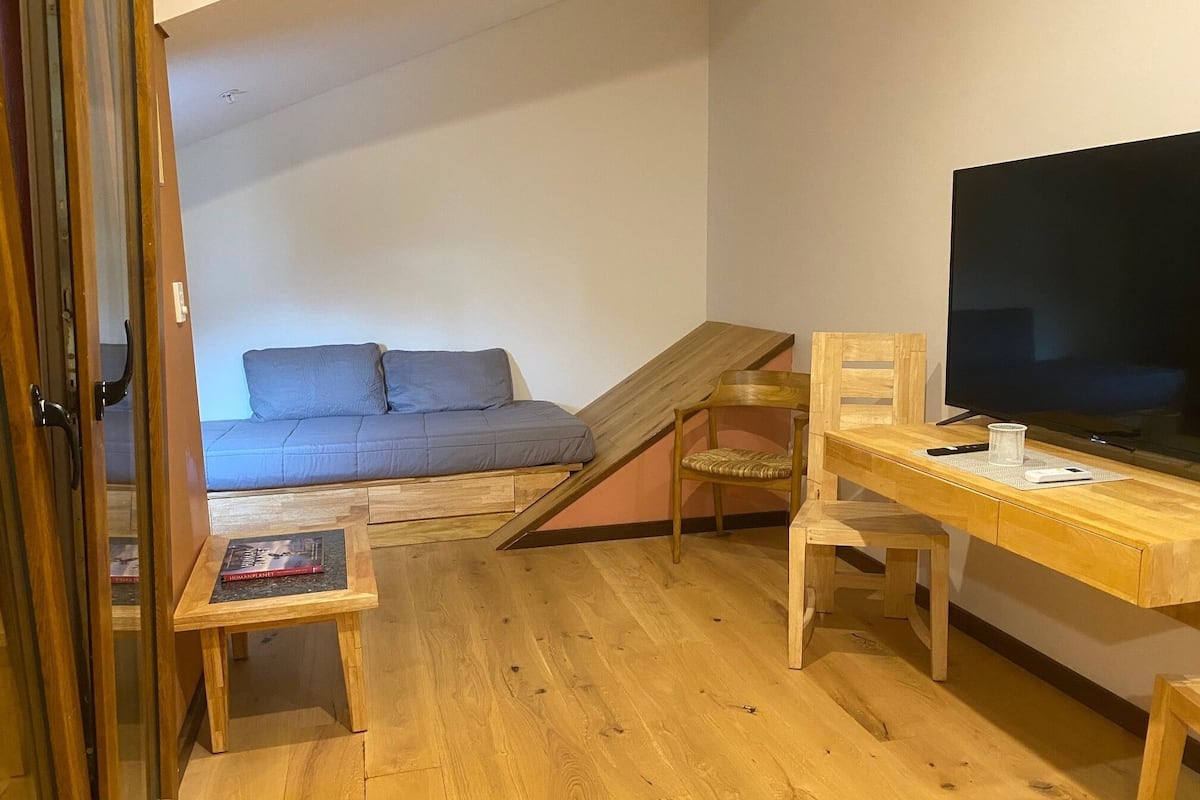 A room with a wooden desk, a television, a sofa, and a small table with books.