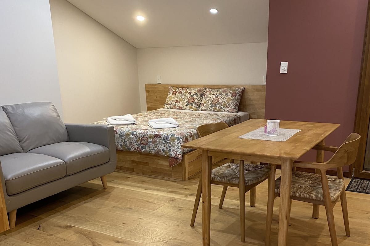 A small, cozy room with a wooden floor, a grey sofa, a wooden table with chairs, and a bed with floral bedding.
