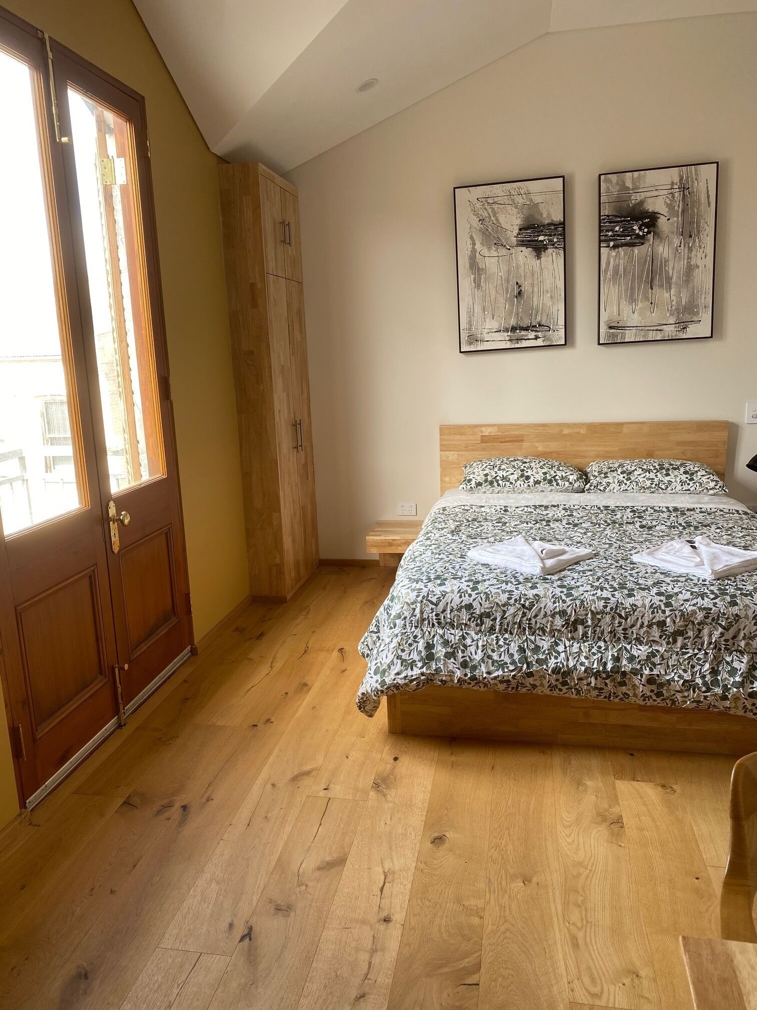 A bedroom with a wooden floor, a bed with floral bedding, a wooden wardrobe, and two abstract paintings on the wall.