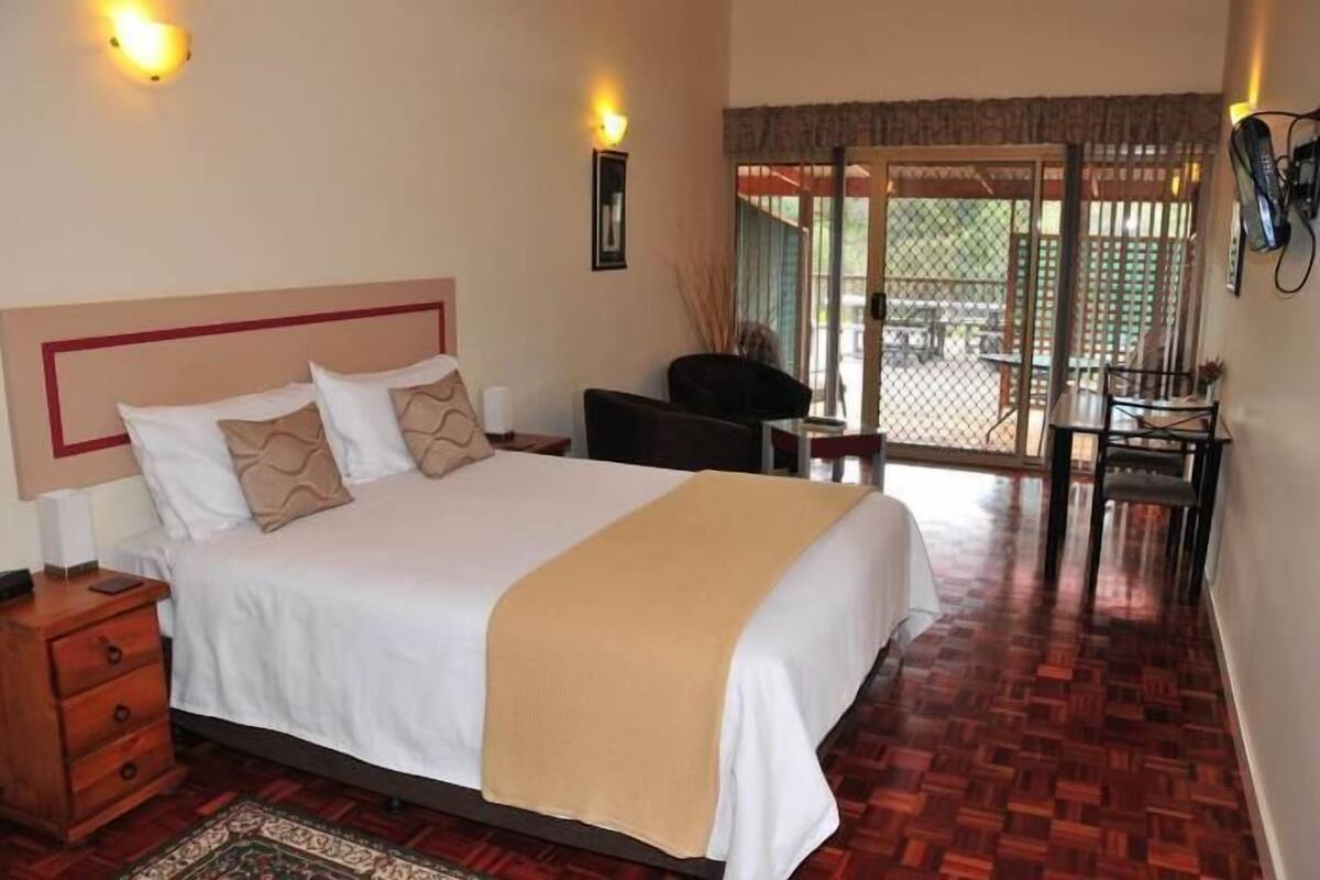 Quarto de hotel com uma cama grande, uma mesinha de cabeceira de madeira, uma escrivaninha com cadeira e vista para área externa através de uma porta de vidro.