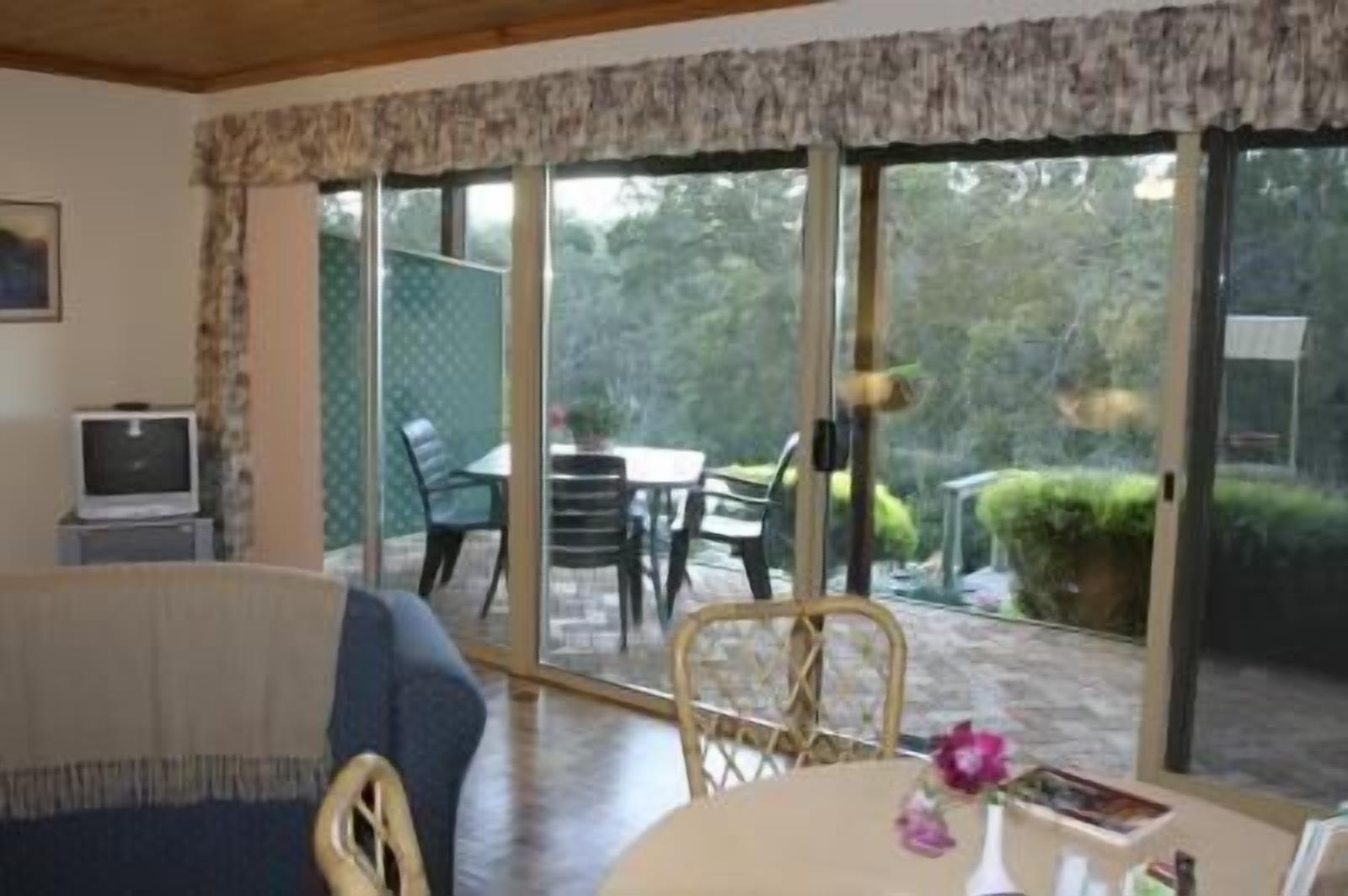 A room with a large sliding glass door leading to an outdoor patio area. Inside, there is a small table with a vase of flowers and a chair.