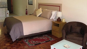 A hotel room with a bed, a nightstand with a lamp, a chair, and a small table. - Tweed Valley Lodge (Bridgetown)