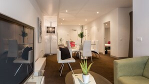 A modern living room with a glass coffee table, a green sofa, and white chairs. There is a TV on the left and a plant on the coffee table. - ABC Accommodation - Flinders (Melbourne)