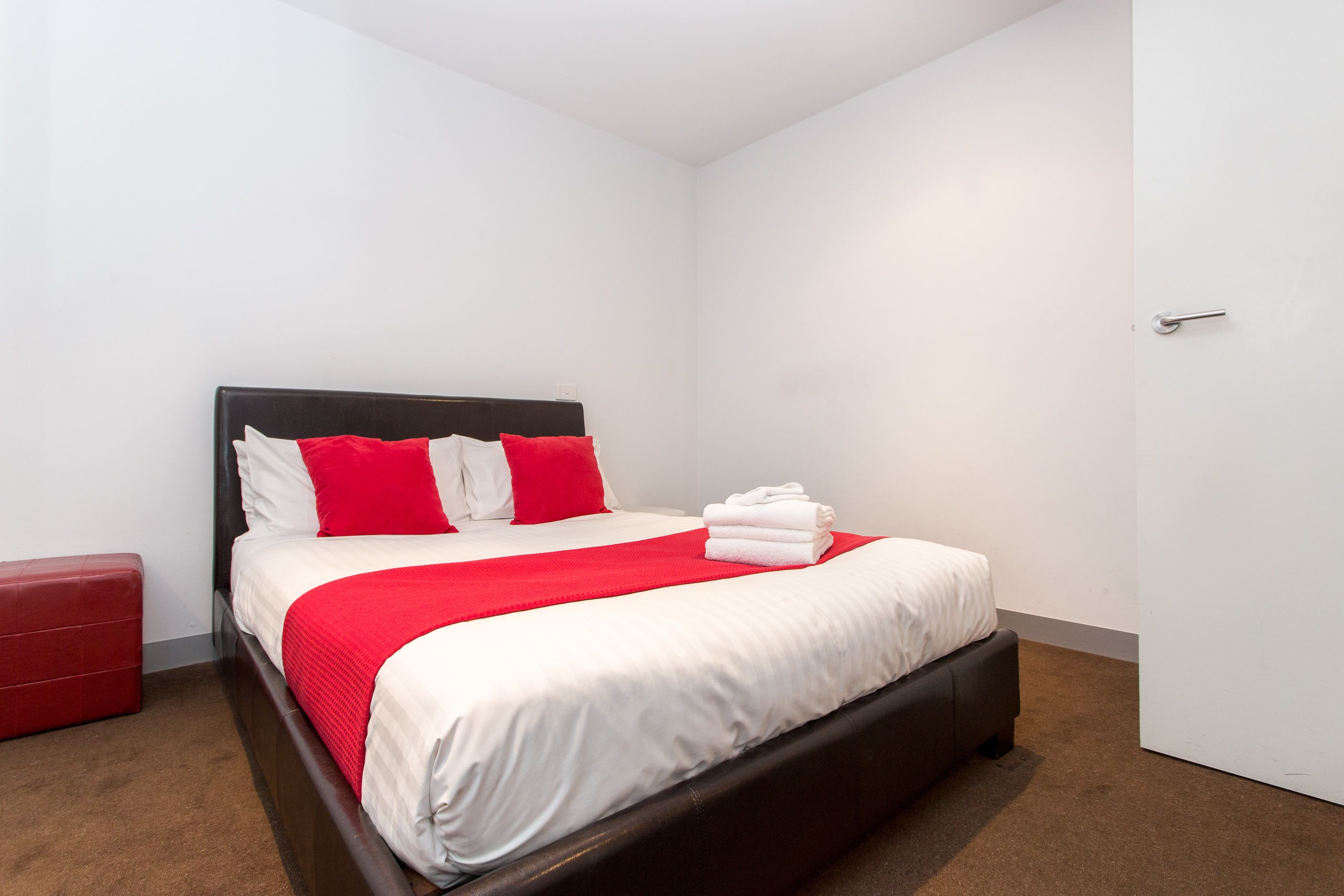 A neatly made bed with white and red bedding, a dark headboard, and a red storage bench.