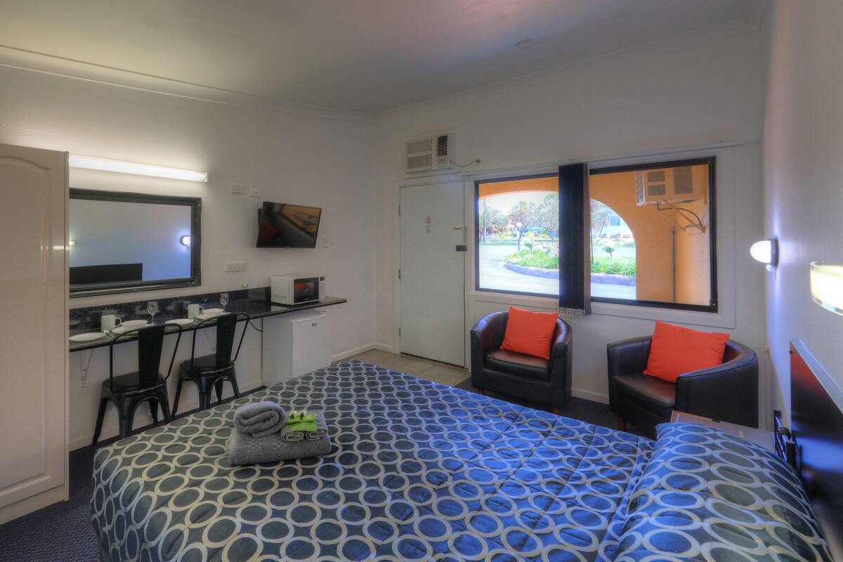 Quarto de hotel pequeno com cama, duas cadeiras, escrivaninha com computador, micro-ondas e uma grande janela com vista para o exterior.