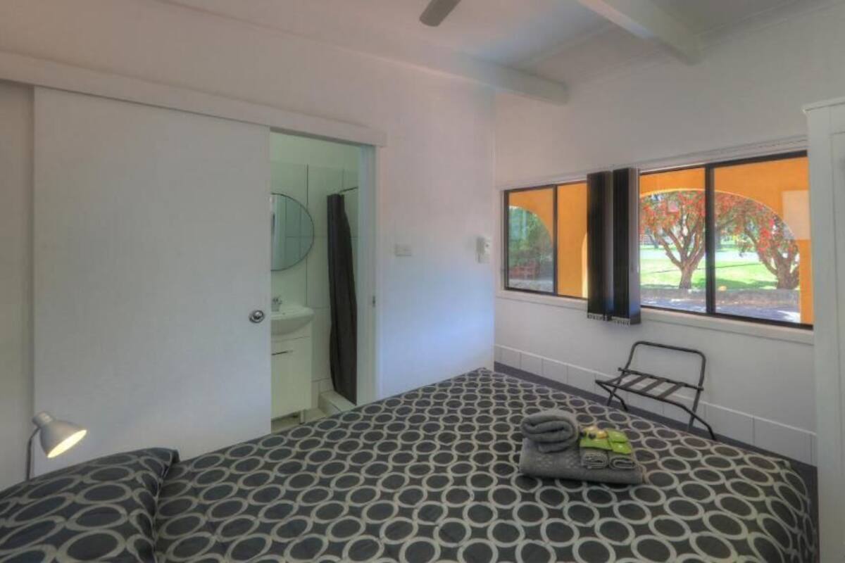 Um quarto com cama, janela com vista para árvores, TV fixada na parede e porta que dá para um banheiro.