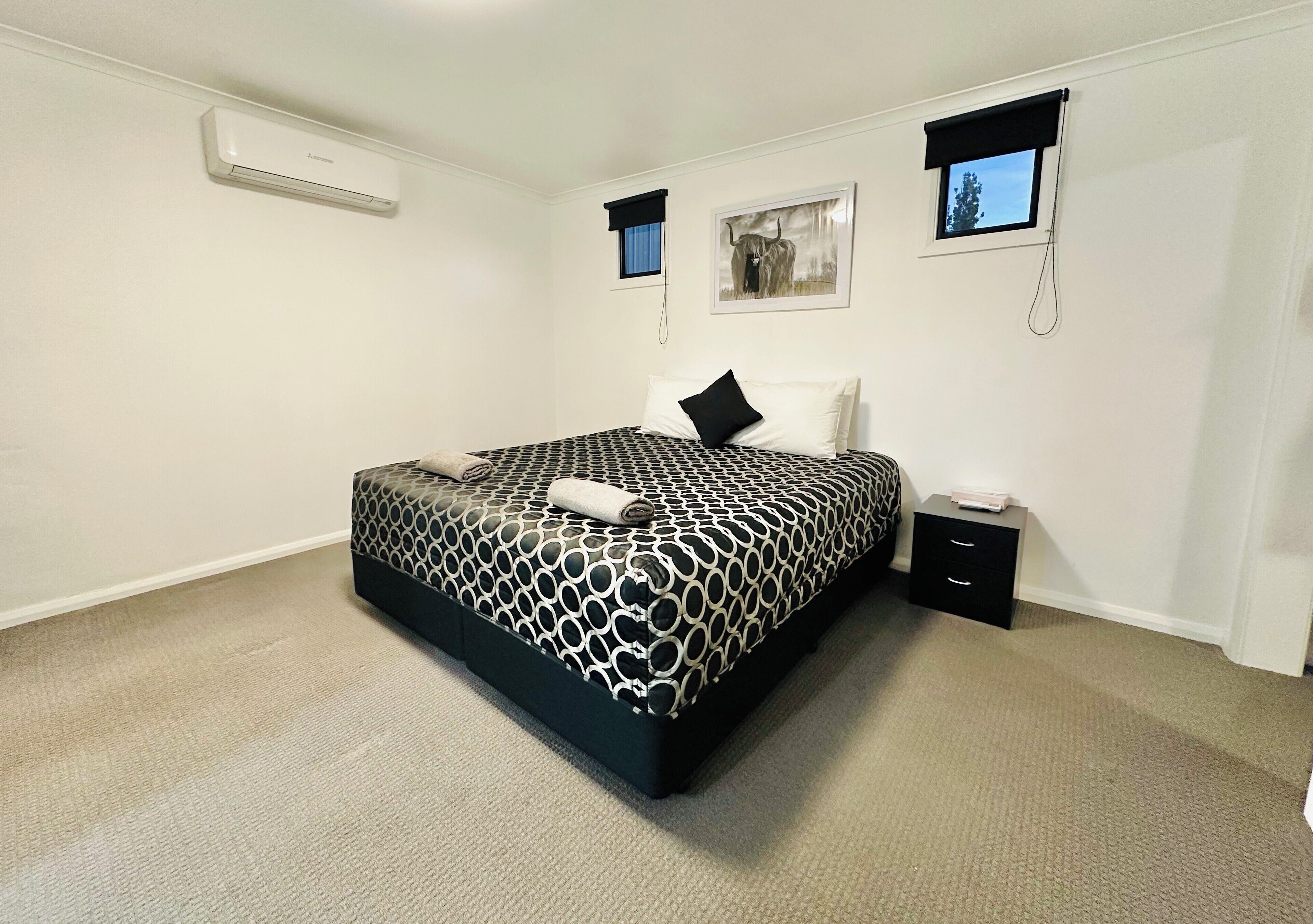 Un dormitorio con cama, mesita de noche, aire acondicionado instalado en la pared y dos ventanas con persianas.
