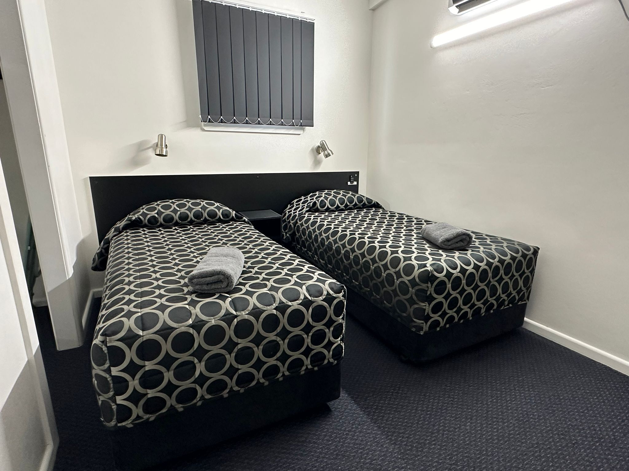 Una habitación con dos camas, cada una con ropa de cama estampada, una ventana con persianas y una mesita entre las camas.