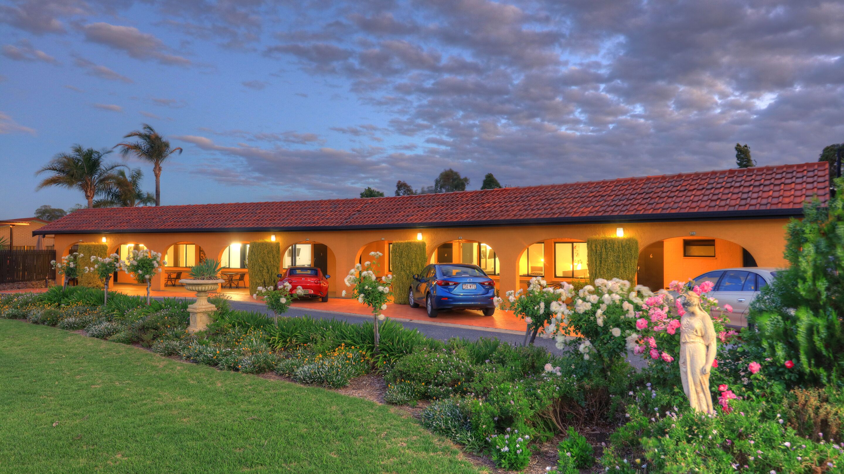 A hotel with a red-tiled roof, multiple parking spaces, and a well-maintained garden with various plants and flowers.